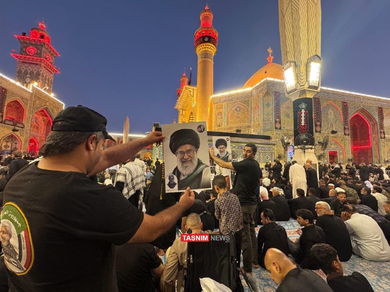 A coffin draped in the Iranian flag is carried above a dense crowd of mourners under red lighting, with flags bearing Persian script held aloft.