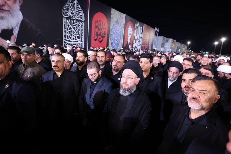 A flag-draped casket bearing green, white, and red stripes is carried above a dense crowd of mourners waving white flags under red lighting.