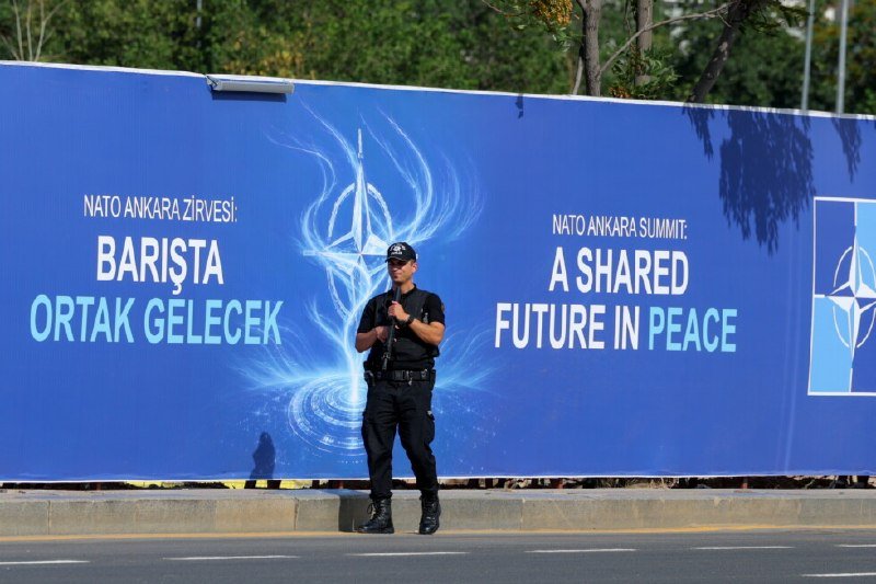 Two men in dark suits sit in black armchairs against a backdrop displaying the NATO logo, "ANKARA" text, and US and Turkish flags, with a small table holding water glasses between them.