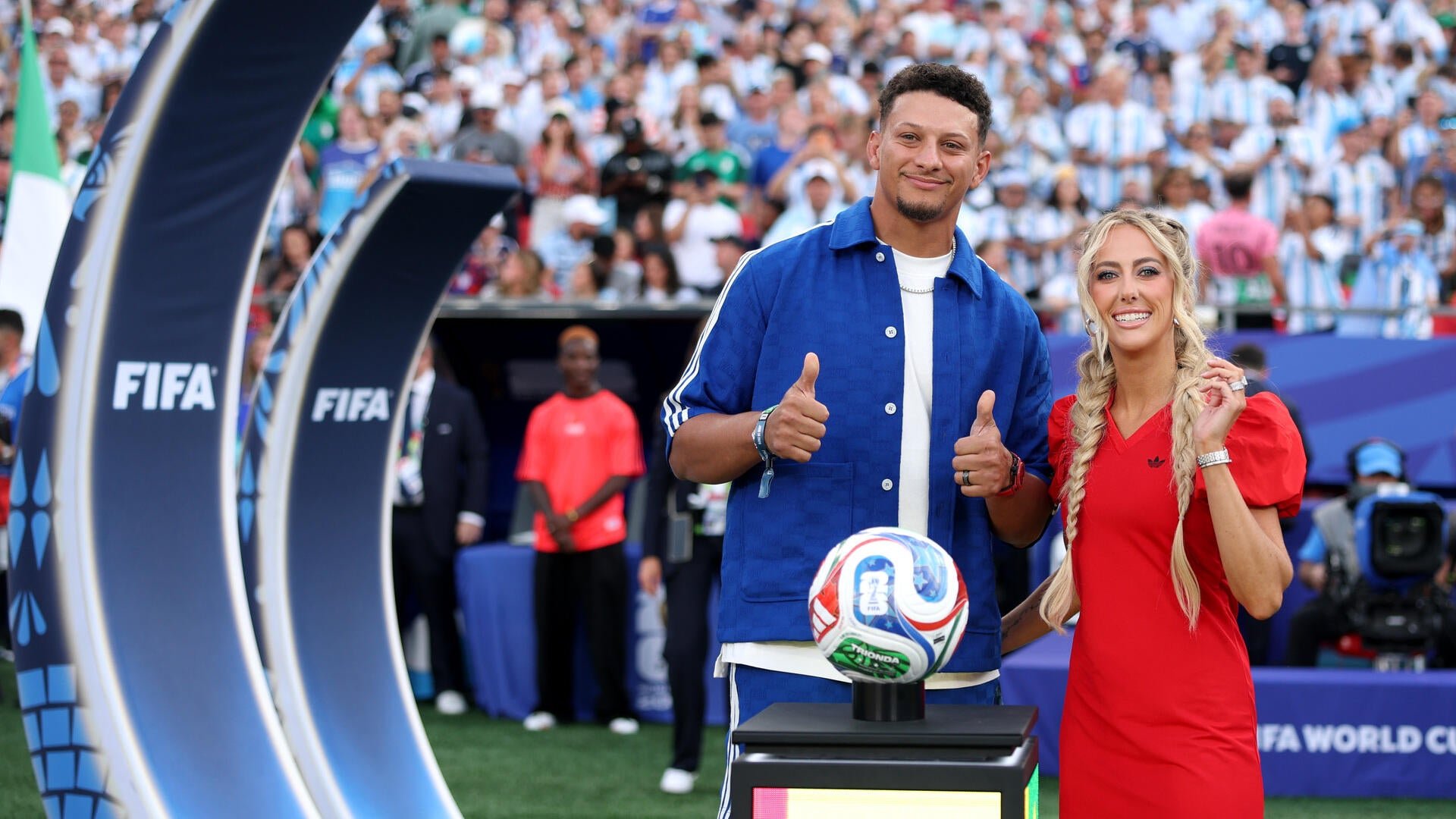 A man in a blue shirt and a woman in a red dress smile and give thumbs up beside a soccer ball on a display, with a packed stadium crowd and FIFA branding visible behind them.