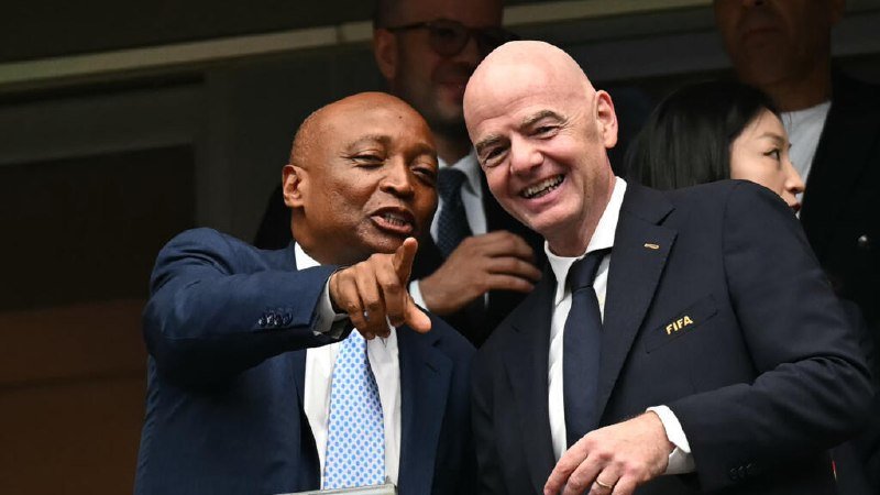 Two men in suits smile and pose together, one pointing forward, with a "FIFA" badge visible on the other man's lapel.