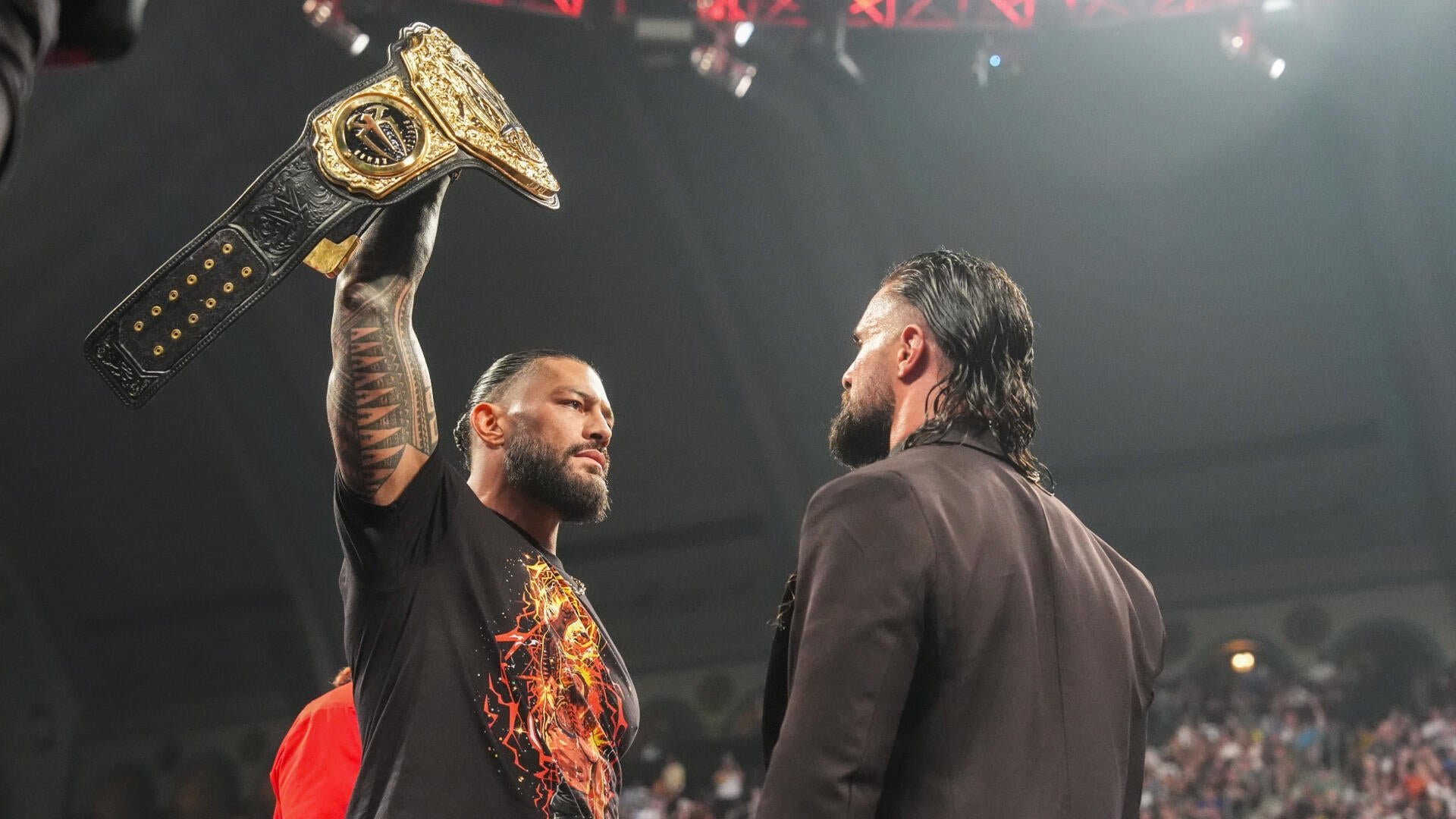 A tattooed man in a black graphic t-shirt raises a championship belt overhead while staring down a long-haired man in a dark jacket during an arena wrestling event.