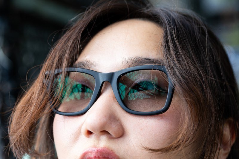 Close-up portrait of a person with brown hair wearing black-framed glasses, with a greenish reflection visible in the lenses.
