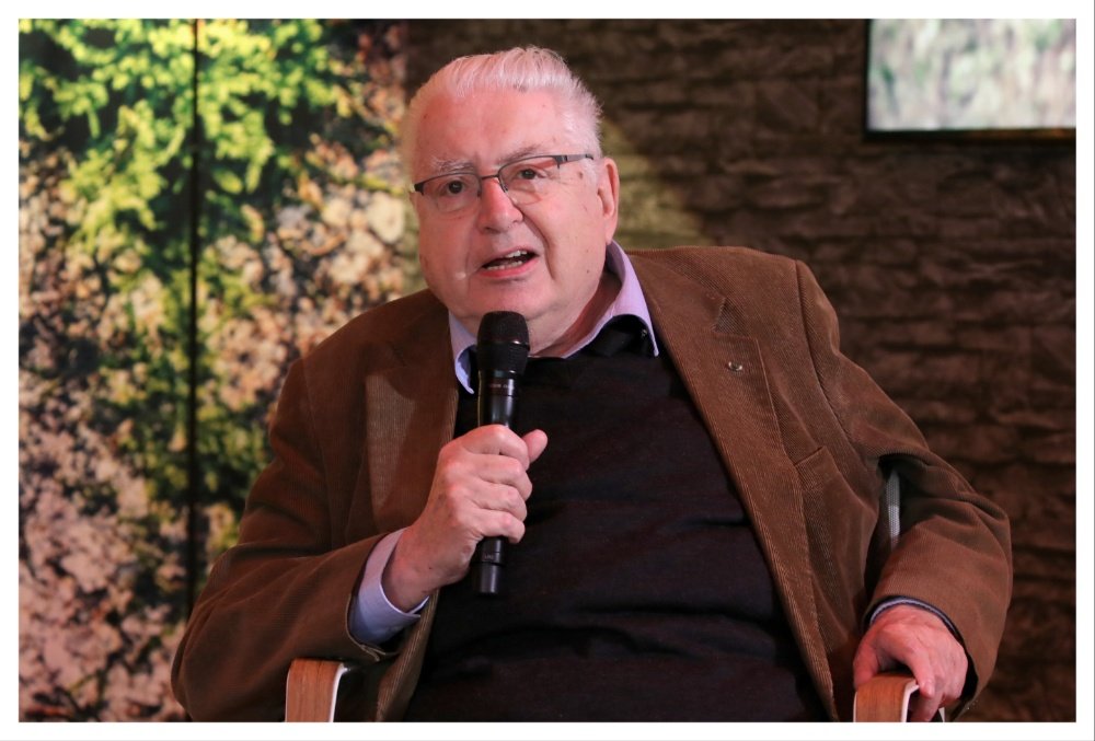 An older man with white hair and glasses speaks into a microphone while seated, wearing a brown jacket over a black sweater, with a floral backdrop and stone wall behind him.