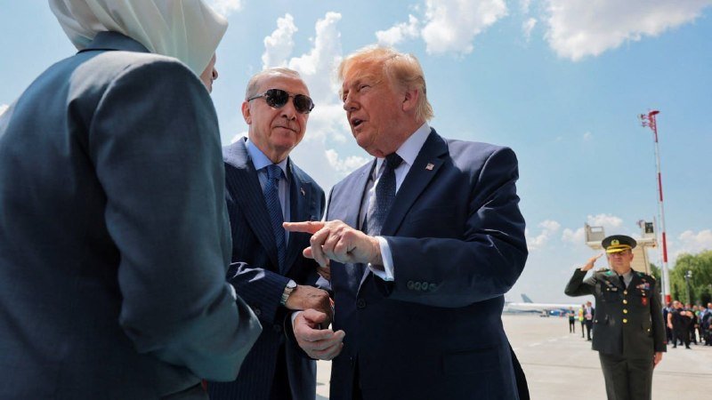 A man in a dark suit and yellow tie speaks with open arms to reporters holding microphones and cameras inside an aircraft cabin featuring a presidential seal.