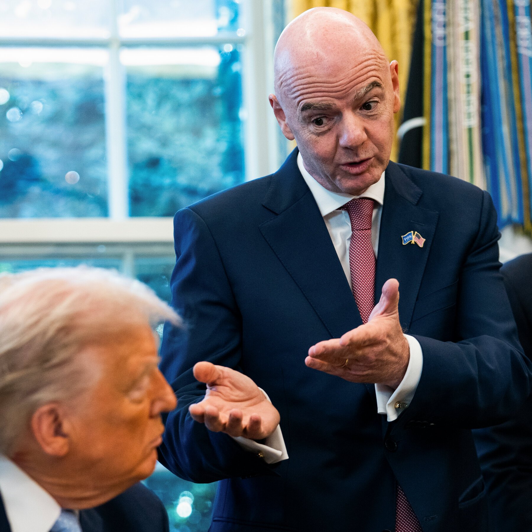Five people sit around a desk in a gold-curtained room with American and military flags, framed portraits, and a model airplane on the coffee table.