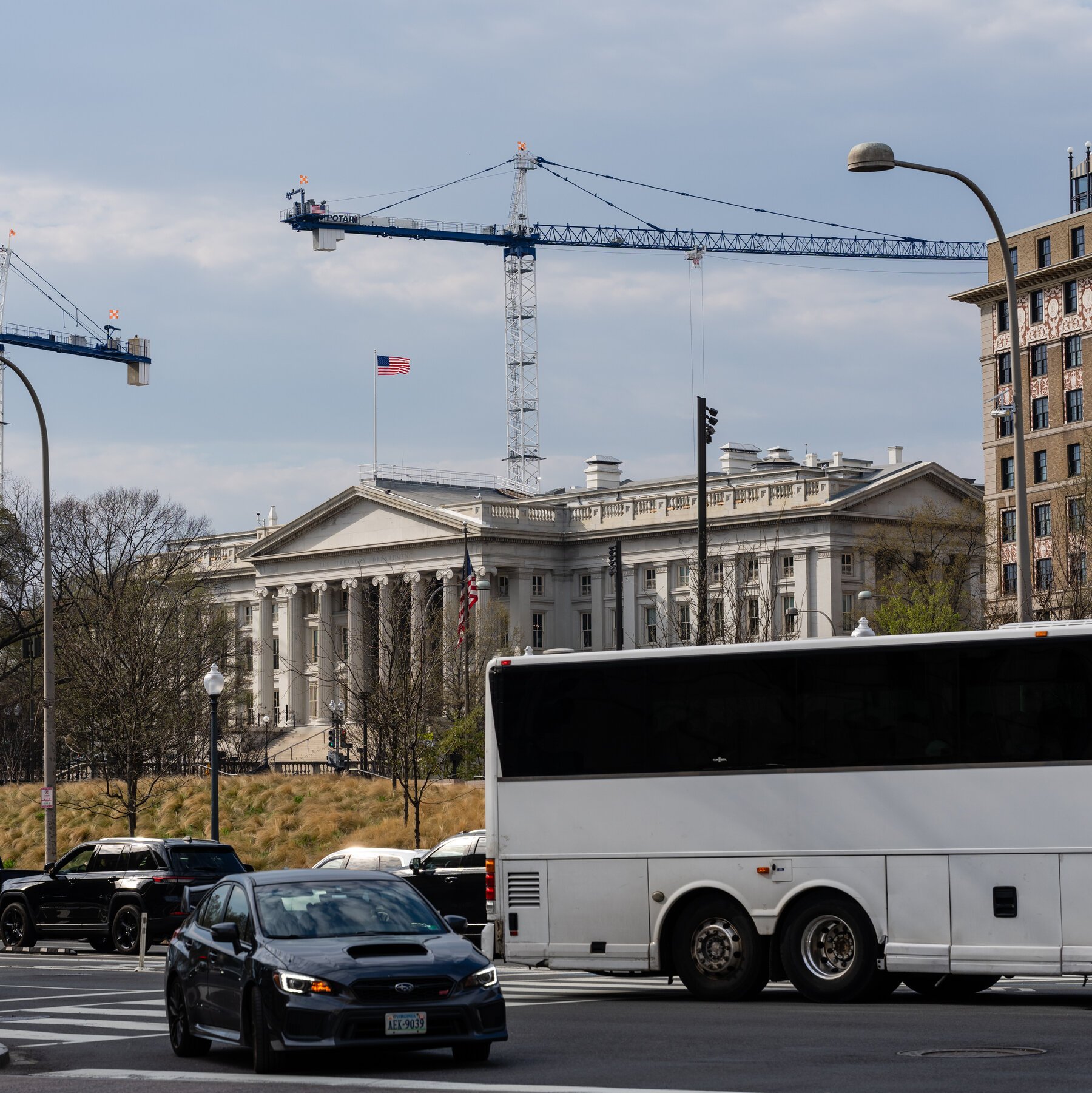 A white bus and a black Subaru drive past a neoclassical stone building with columns flying an American flag, while a construction crane towers overhead.