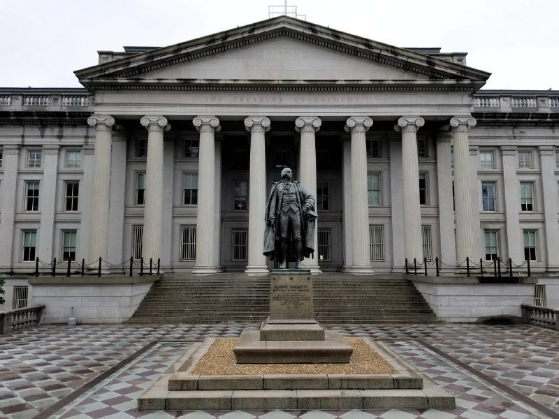 A bronze statue of a man stands on a pedestal in front of a large neoclassical building inscribed "THE TREASURY DEPARTMENT," featuring tall columns and a triangular pediment.