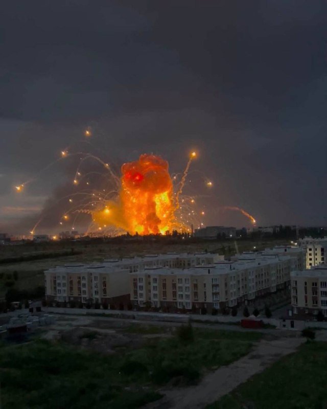 A large orange explosion erupts near a row of residential apartment buildings at dusk, with bright streaks arcing outward into the dark sky.