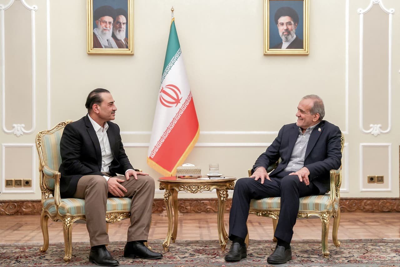 Two men in suits sit facing each other in ornate chairs in a formal room, with an Iranian flag and two framed portraits hanging on the wall behind them.
