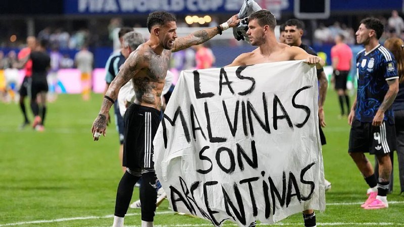 Two shirtless soccer players on a stadium field hold a white banner reading "LAS MALVINAS SON ARGENTINAS" after a match, with a referee and teammates visible nearby.