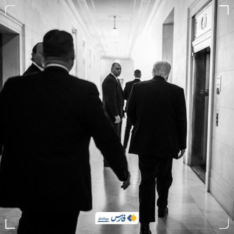 Black-and-white photograph of several men in dark suits walking down a formal hallway toward an elevator, viewed from behind.