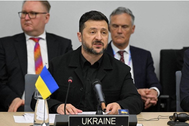 A man in a black shirt speaks at a microphone behind a "UKRAINE" placard, with a small Ukrainian flag beside him, as two men in suits sit behind.