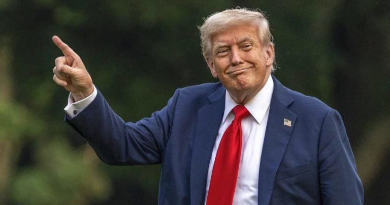 A man with light hair wearing a navy blue suit, white shirt, and red tie points upward with his right hand while smiling, set against a blurred green background.
