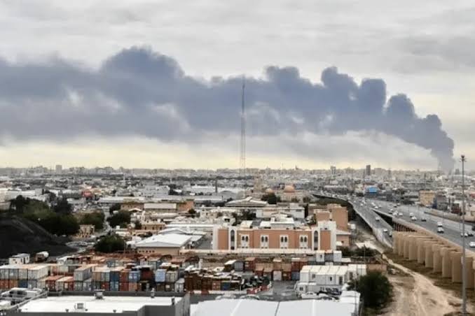 A wide shot shows a sprawling urban area with shipping containers in the foreground, low-rise buildings, a highway, and a tall communications tower beneath a cloudy sky.