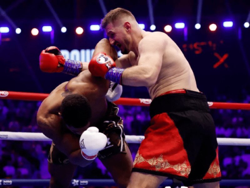 A boxer in black-and-gold trunks ducks under a punch from an opponent in red-and-black trunks during a professional boxing match inside a brightly lit ring.