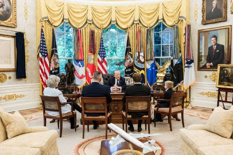 Four people sit around a desk in the Oval Office, facing a man seated behind it, surrounded by flags and framed portraits.
