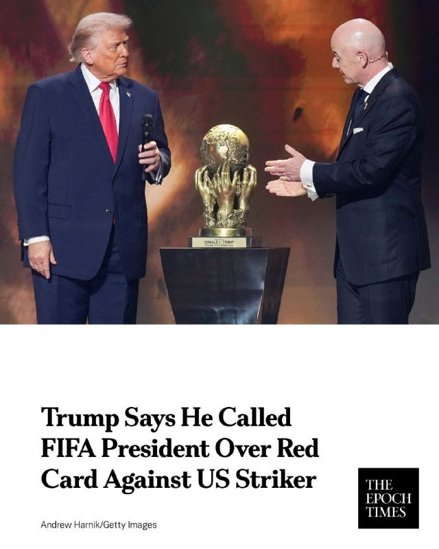 A bald man in a dark suit and tie smiles while taking a selfie with a smartphone, standing beside a taller man with blonde hair in a dark suit and red tie.