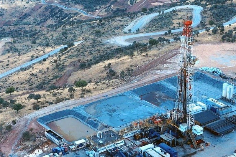 An aerial view shows an oil drilling rig, lined containment ponds, storage tanks, and modular buildings situated in a dry, hilly landscape with winding dirt roads.