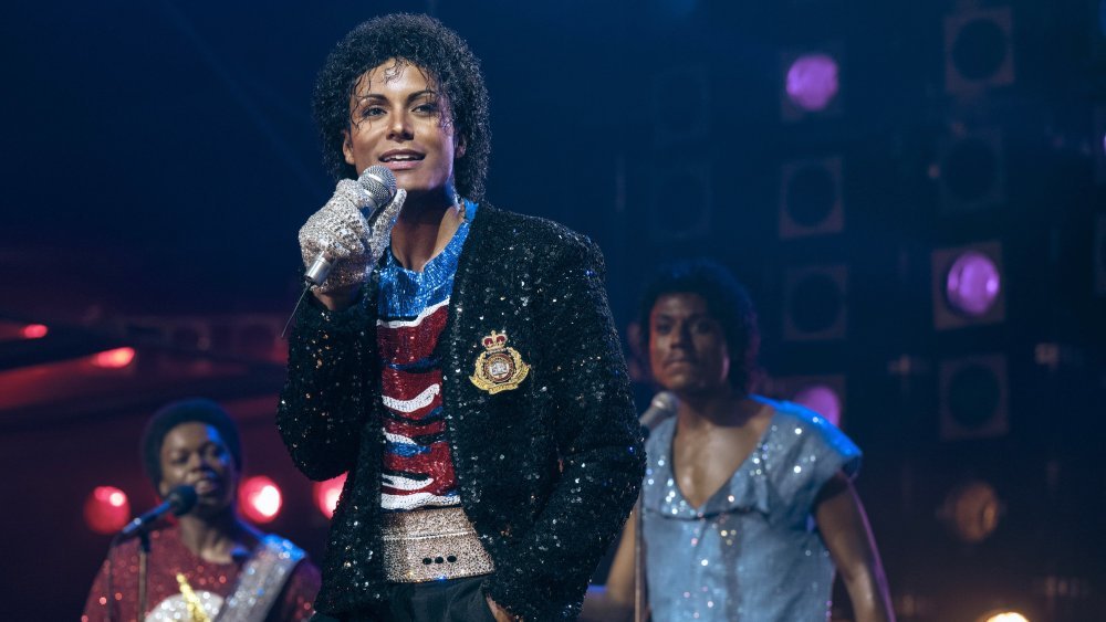 A man with curly hair sings into a microphone on a stage, wearing a sequined black jacket over a red, white, and blue top, flanked by two backup singers.