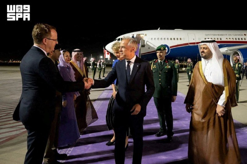 Two men shake hands on a purple carpet at night in front of a Government of Canada aircraft, surrounded by officials and military personnel.