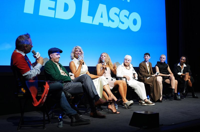 Eight people sit on a stage in chairs, some holding microphones, in front of a large screen displaying "TED LASSO."