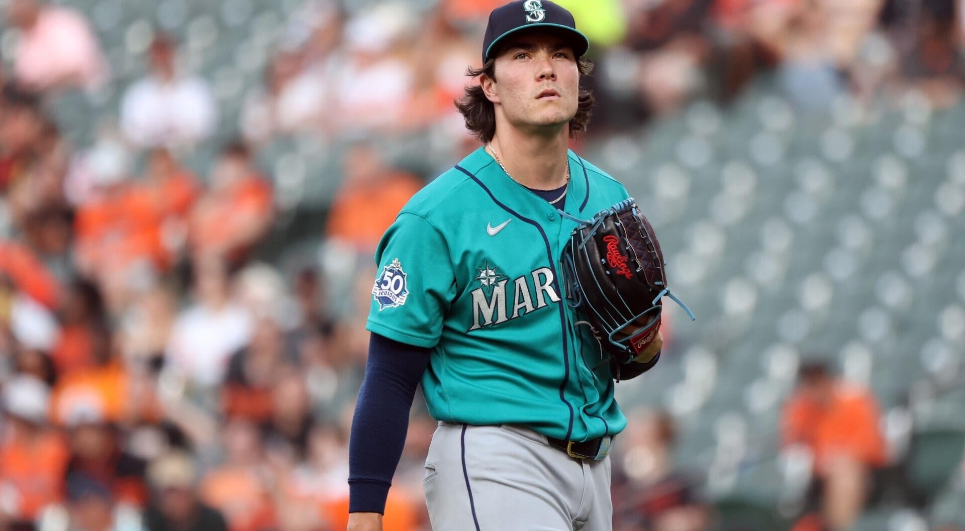 A baseball pitcher in a teal Mariners jersey and cap stands on the field, holding a glove and looking upward during a game.