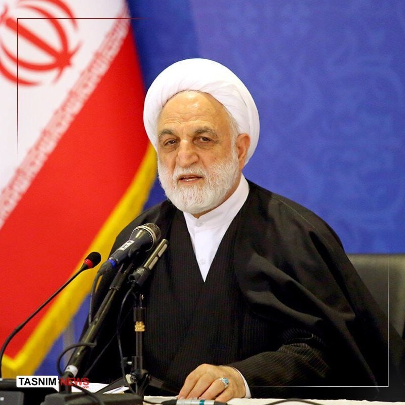 A white-bearded man wearing a white turban and black clerical robes speaks at a microphone, with an Iranian flag visible behind him.