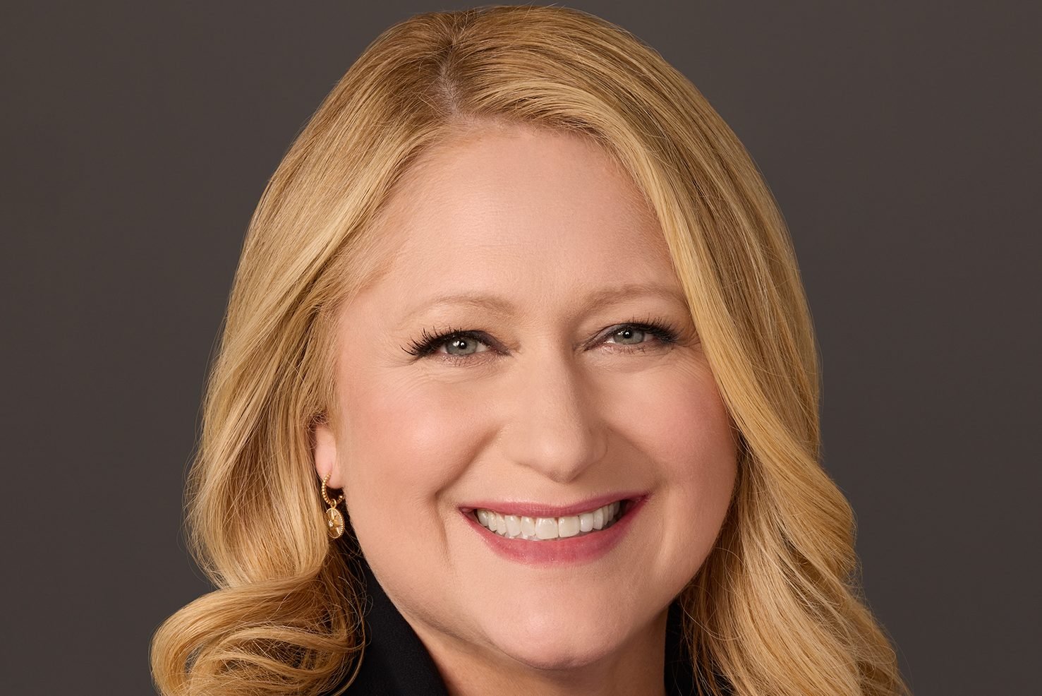 A smiling woman with long blonde hair and gold hoop earrings poses for a portrait against a dark gray background.