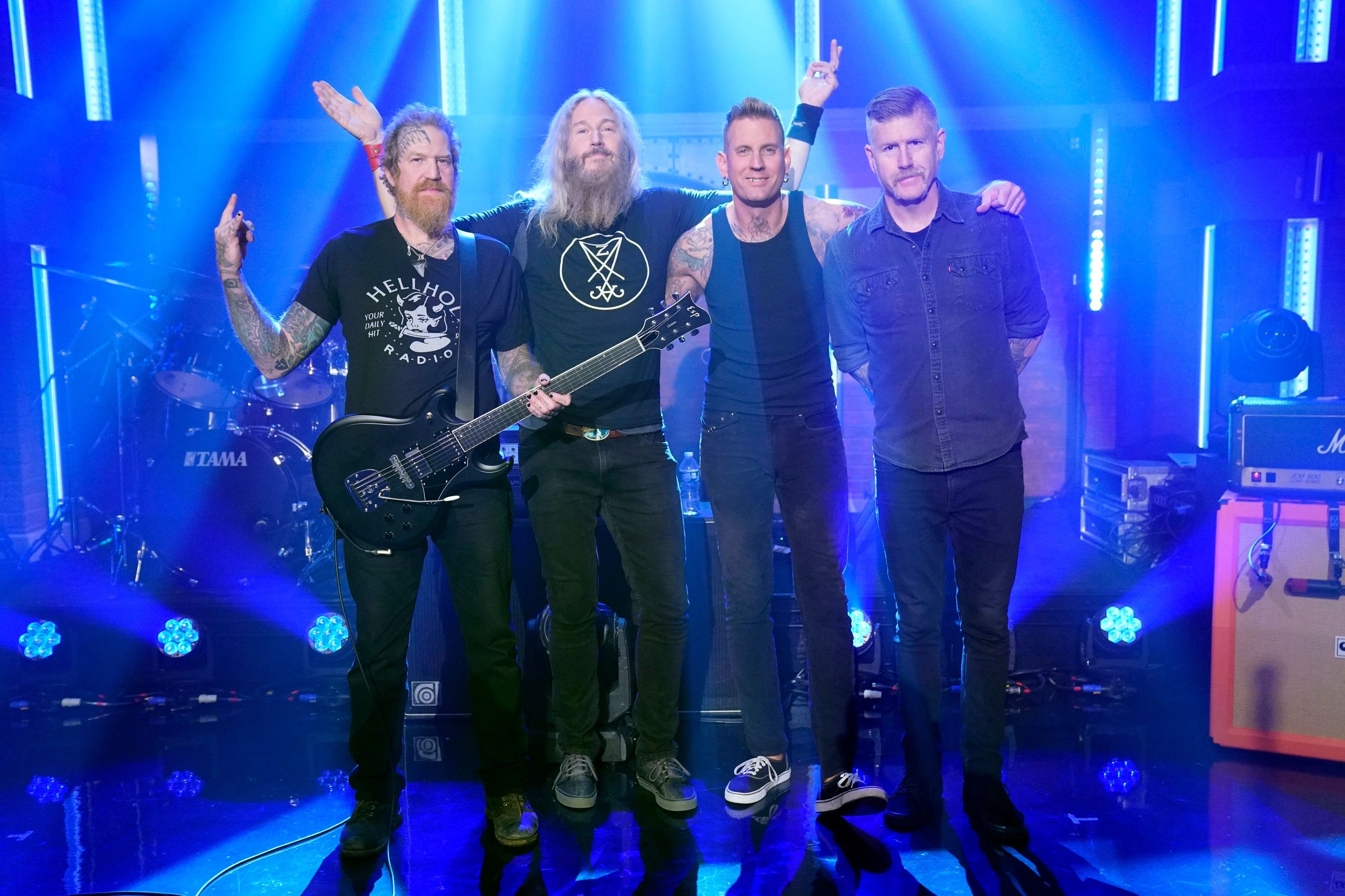 Four tattooed men pose together on a stage illuminated by blue lighting, with one holding a black electric guitar, a TAMA drum kit, and amplifiers visible behind them.
