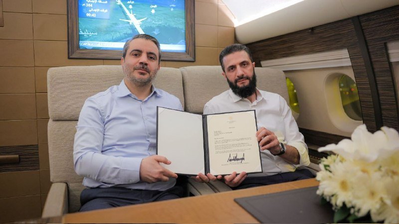 Two bearded men sit inside a luxury aircraft cabin, jointly displaying an open, signed document folder toward the camera.