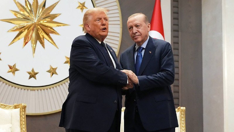 Two men in dark suits shake hands in front of a decorative emblem featuring gold stars and a red-and-white flag.