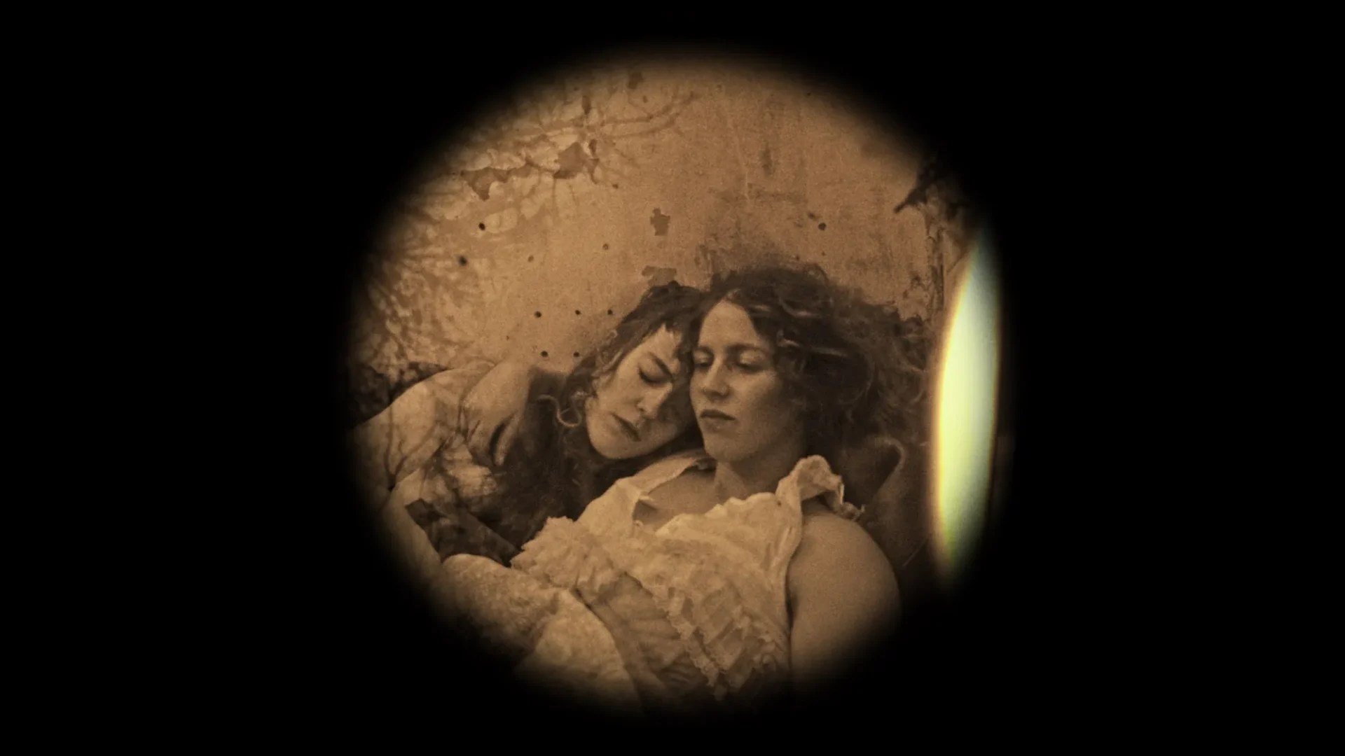 Viewed through a circular vignette, two women with closed eyes rest their heads together against a textured backdrop in a sepia-toned photograph.