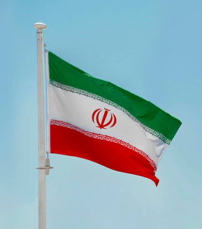 The national flag of Iran—featuring green, white, and red horizontal stripes with a central red emblem—waves from a pole against a clear blue sky.