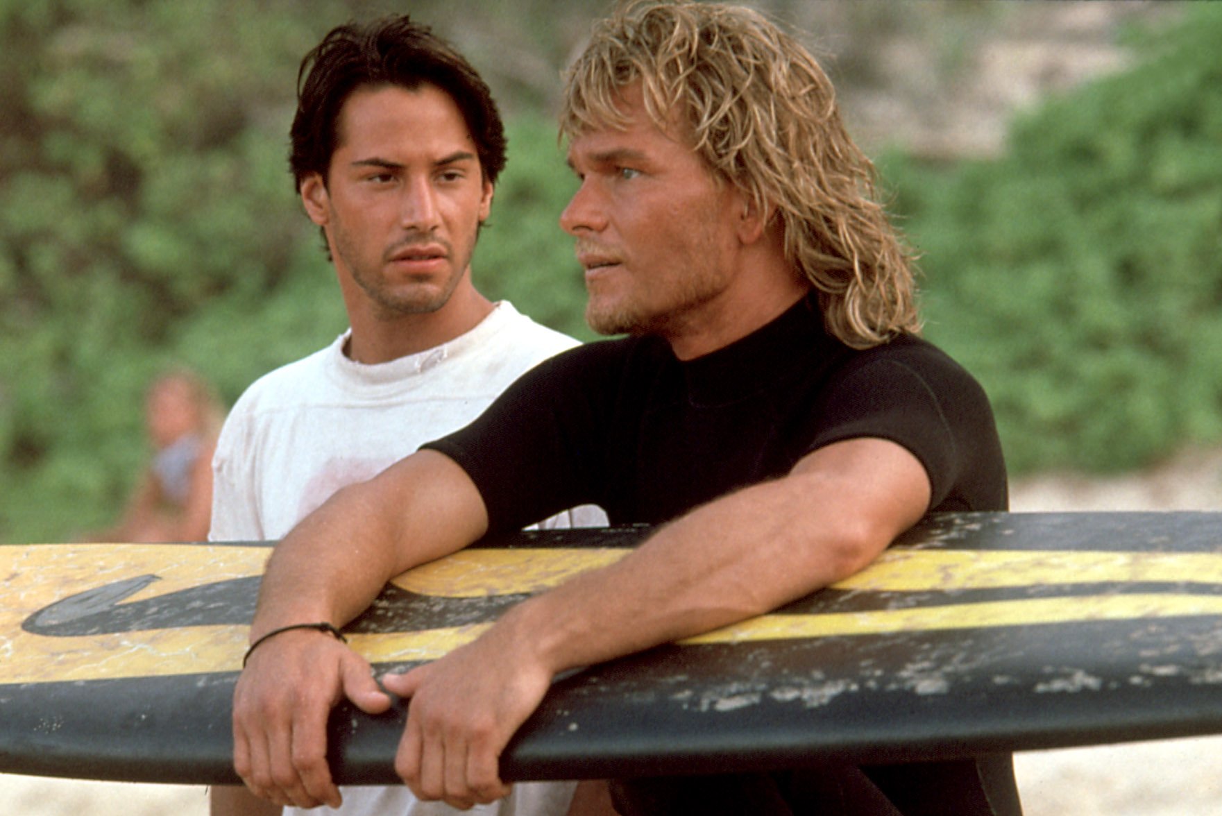 Two men carrying surfboards walk along a sunlit beach with green foliage in the background.
