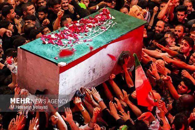 A dense crowd carries a coffin draped in a green, white, and red flag with Arabic script, topped with scattered rose petals and a green flag nearby.