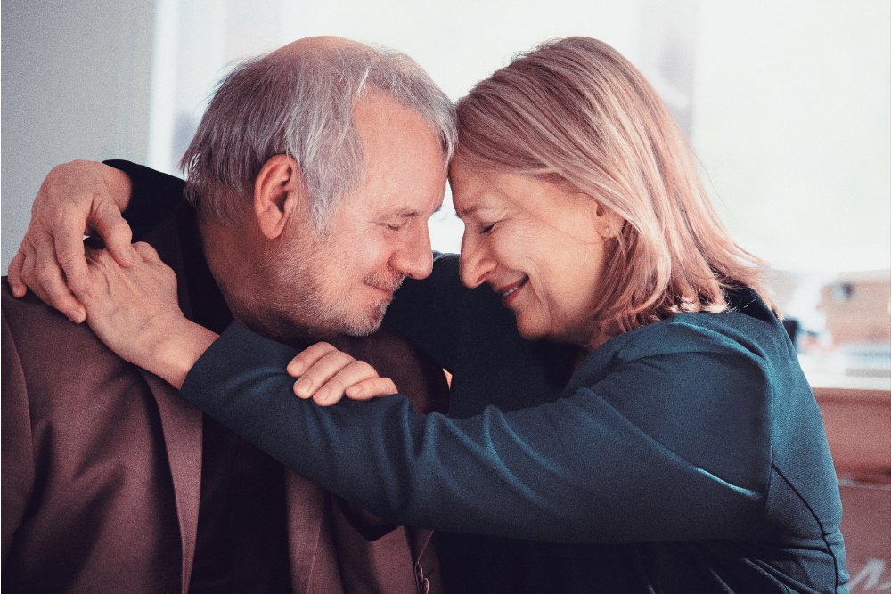 A smiling gray-haired man and blonde woman embrace forehead-to-forehead indoors, her arm draped over his shoulder.