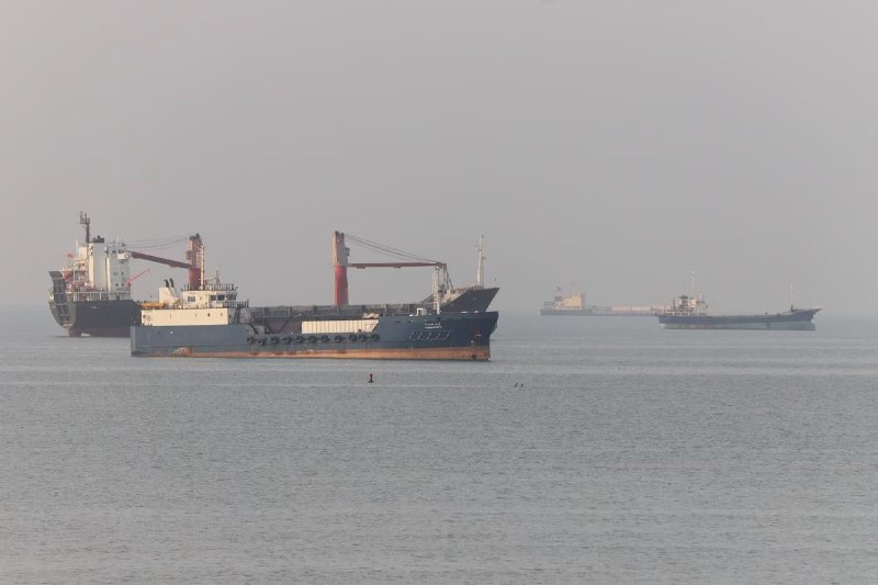 A large cargo ship named "AMERICAN FREEDOM" from Wilmington, DE sits in a port alongside a tugboat, with stacked shipping containers and cranes visible in the background.
