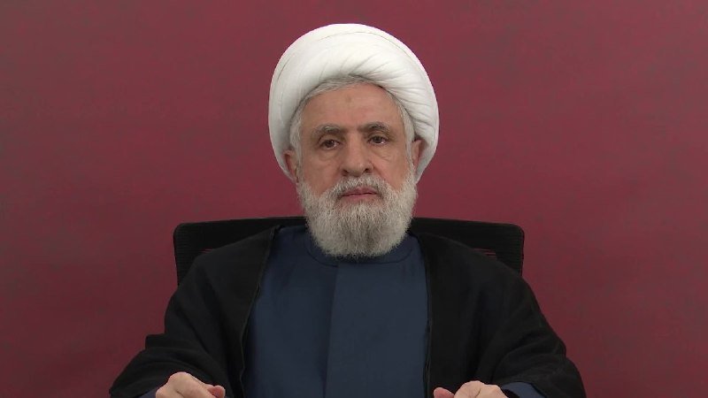 A bearded man wearing a white turban and dark robes sits against a maroon background, gesturing with his hands.