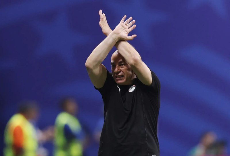 A man in a black athletic shirt stands with his hands raised above his head, clapping, against a blue stadium backdrop.