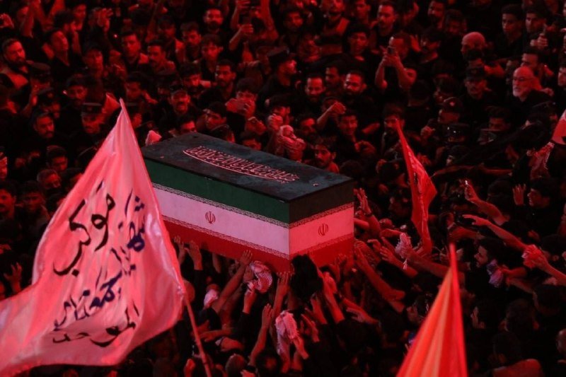 A flag-draped coffin bearing the Iranian flag colors is carried through a dense, red-lit crowd of mourners with raised hands.