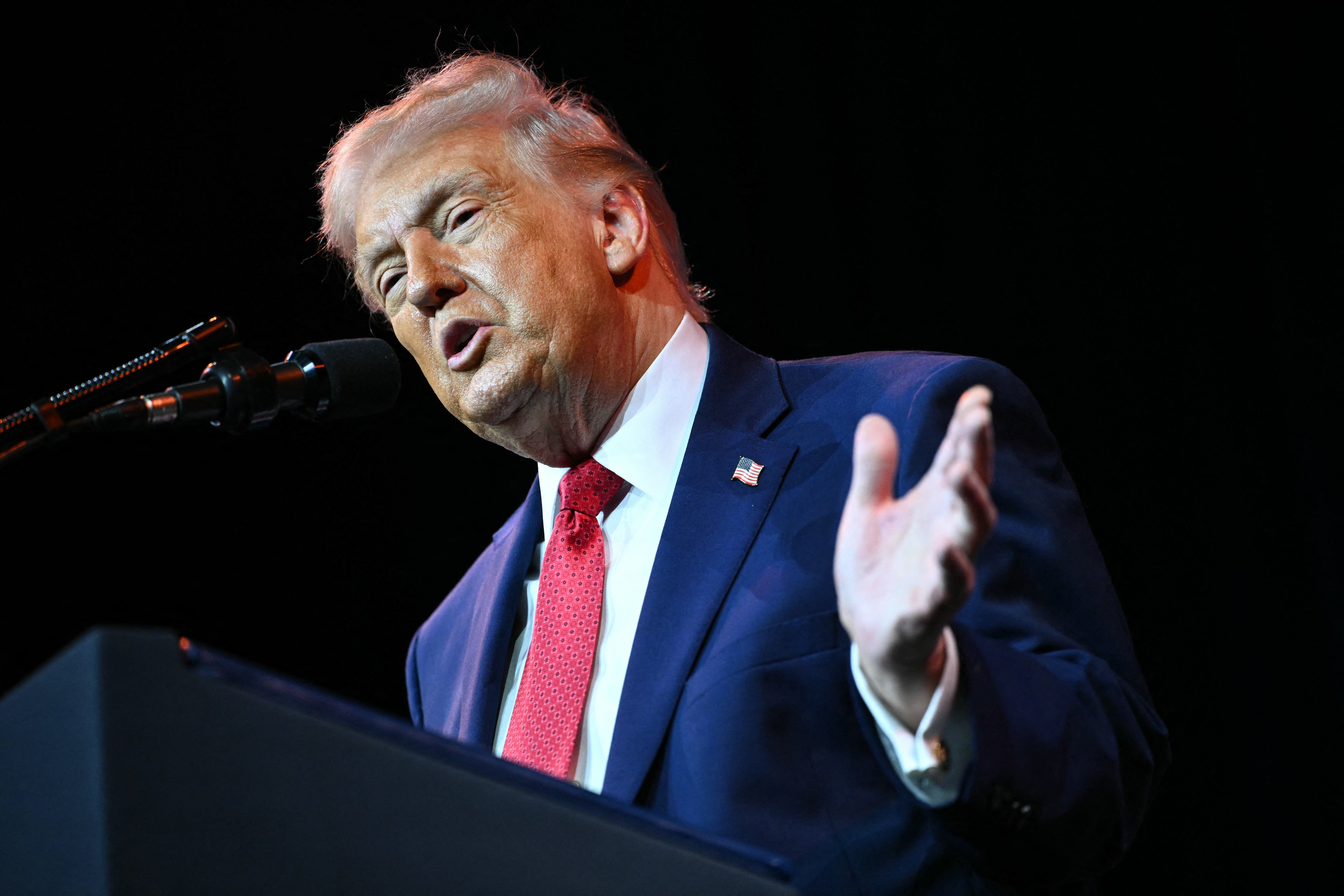 An older man with white hair, wearing a navy suit, white shirt, red tie, and American flag lapel pin, speaks at a podium with microphones against a black background.
