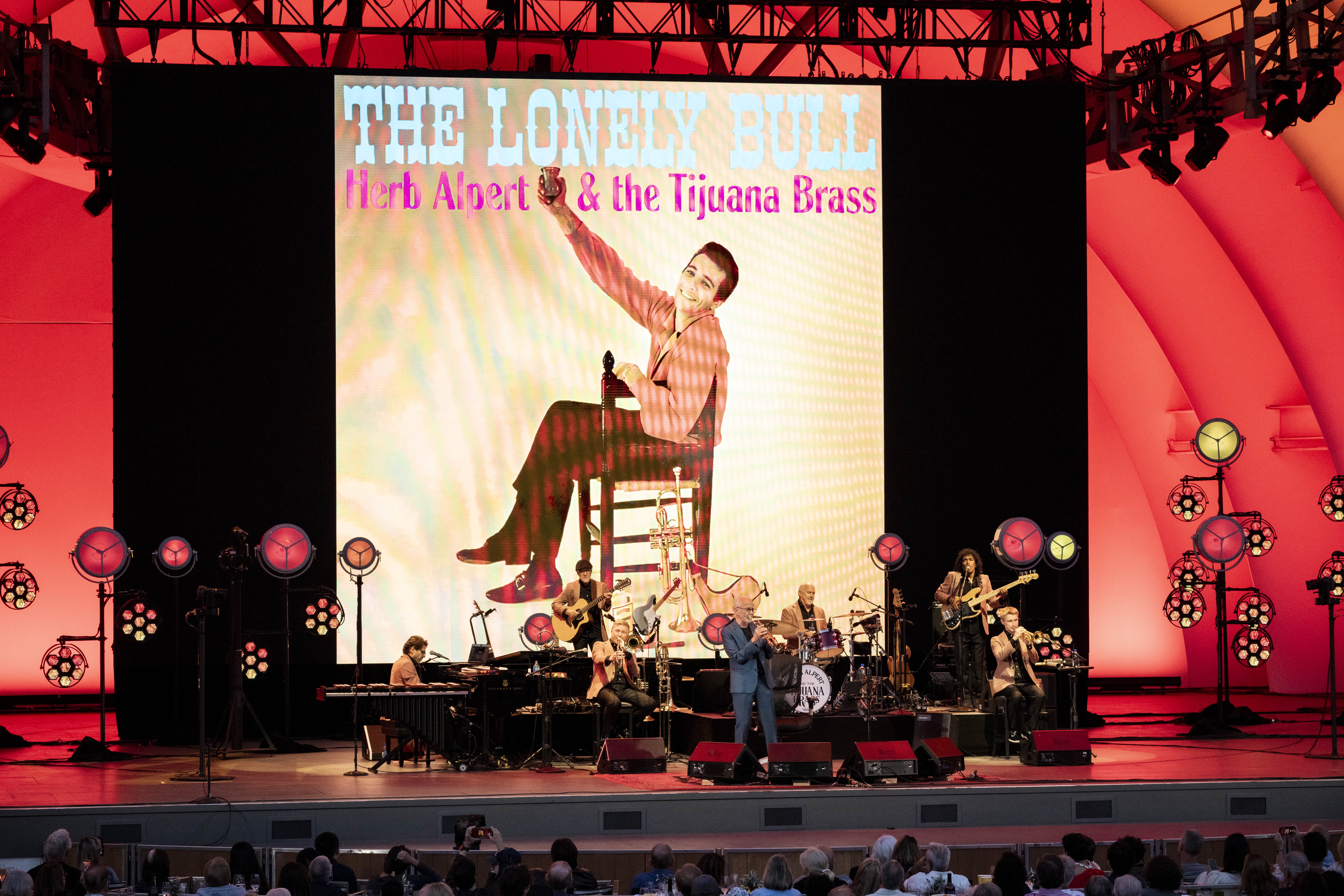 A band performs on a red-lit stage with musicians playing trumpet, guitar, bass, drums, and keyboards before a large screen displaying "The Lonely Bull Herb Alpert & the Tijuana Brass" album artwork.