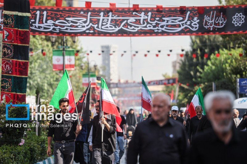 People wearing black walk down a street carrying Iranian flags and black banners beneath an overhead black banner displaying Arabic script.
