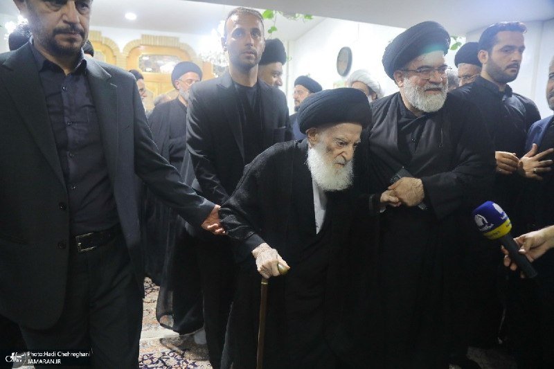 A white-bearded elderly cleric in black robes and turban walks with a cane, supported by men in dark suits, as a reporter holds a microphone nearby.