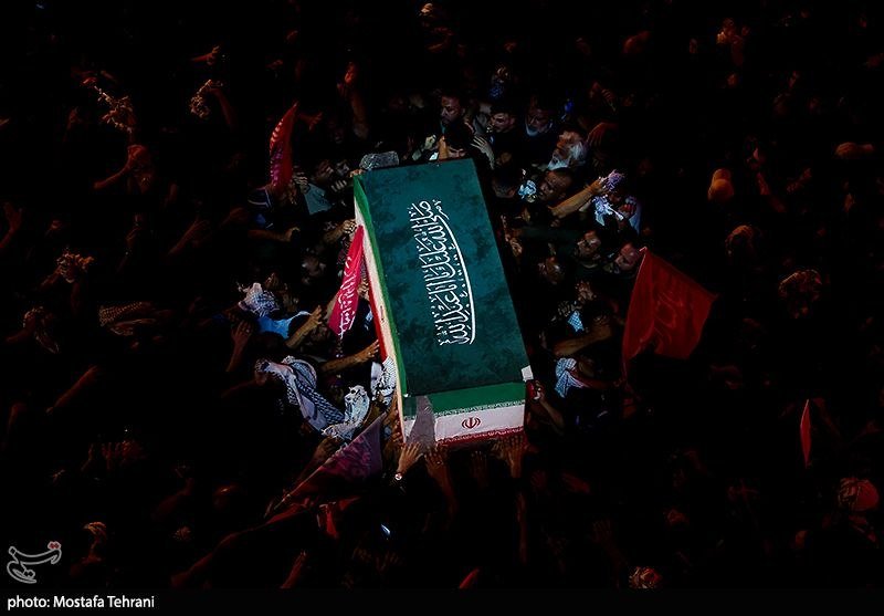 A green-draped casket bearing Arabic script is carried through a dense crowd at night, surrounded by raised hands and red and black flags.