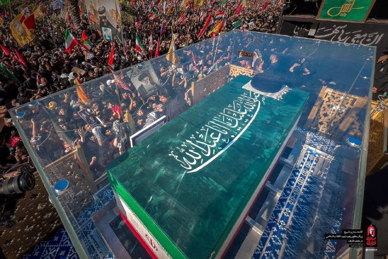 A large crowd carrying flags and banners gathers behind a transparent enclosure holding a green-covered casket inscribed with Arabic calligraphy.