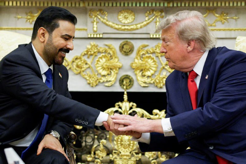 Two men in dark suits—one wearing a blue tie, the other a red tie—shake hands while seated in front of an ornate gilded decorative backdrop.