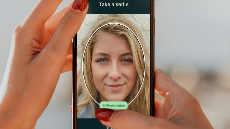 Two hands with red nail polish hold a smartphone displaying a blonde woman's face within an oval frame, with "Take a selfie" at the top and a green "Photo taken" banner at the bottom.