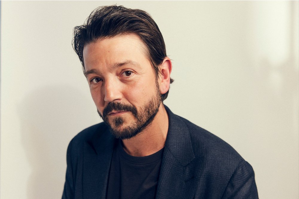 A bearded man with dark hair, wearing a dark blazer over a black t-shirt, looks directly at the camera against a plain white background.
